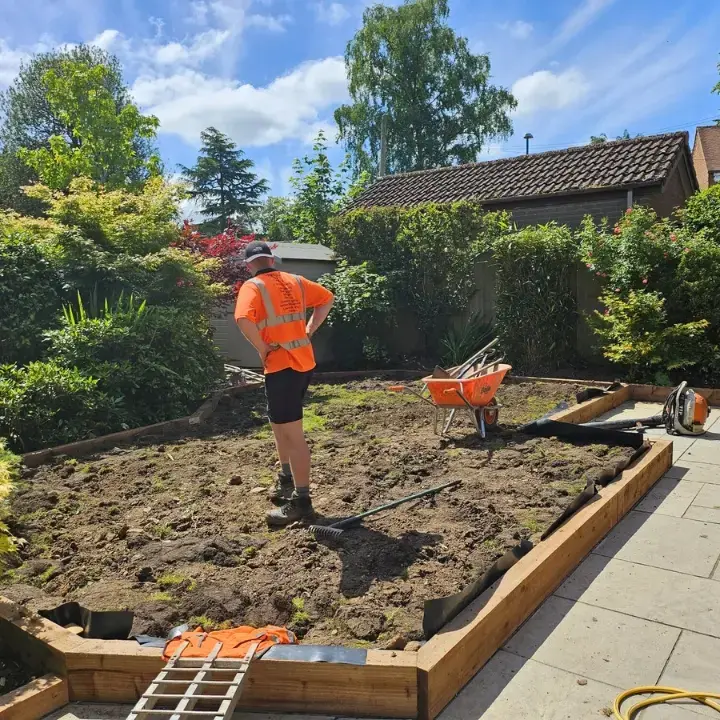 Outdoor maintenance operative working in established garden on lawn levelling with sleeper borders set in place around the lawn and patio areas.